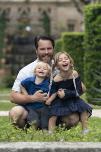 A man kneels in a garden, smiling with a young boy and girl, both children laughing and facing the camera—capturing a joyful moment of family photography Miami style.