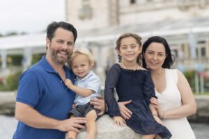 A family of four poses outdoors by the water; the father and young son stand on the left, while the mother and daughter sit on a stone ledge on the right, capturing a perfect moment of family photography Miami style.