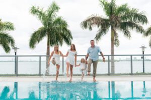 Four people, two adults and two children, walk barefoot by a pool, splashing water. Palm trees and a body of water are visible in the background under a cloudy sky—perfect for family photography in Miami.