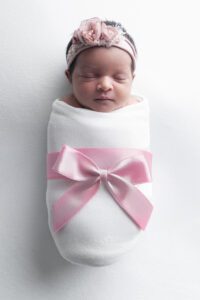 Newborn baby wrapped in a white blanket with a pink satin bow, wearing a pink floral headband, lying on a white background—captured by a Miami newborn photographer.