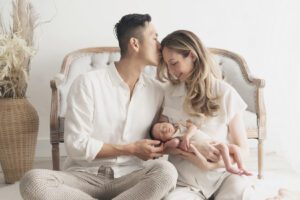 A Miami newborn photographer captures the precious moment of a man and woman holding a baby in a white chair.