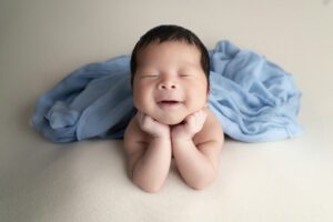 A newborn baby with closed eyes and a smile rests chin on hands, lying on a light surface with a blue cloth draped behind—captured by a Miami newborn photographer.