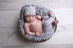 A Miami newborn photographer captures a serene moment as a newborn baby sleeps in a wooden bowl, wearing a grey hat and matching pants, lying on a fluffy grey blanket.