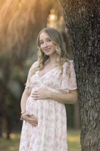 Pregnant woman in a floral dress stands outdoors, leaning against a tree, with hands resting on her belly and smiling at the camera.