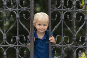 Young child with blond hair, wearing a blue shirt, stands behind ornate iron gate, looking through the bars with a slight smile. Green foliage is visible in the background.