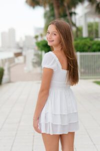 A teenage girl with long brown hair wearing a short white dress stands outdoors on a paved walkway, looking back over her shoulder and smiling.