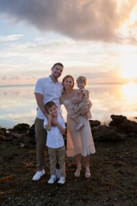 A family of four stands together near a rocky shoreline at sunset, smiling at the camera with calm water and a cloudy sky in the background—a perfect moment of family photography in Miami.