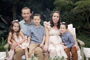 A family of five sits on a bench outdoors, captured in this family photography Miami moment. Two adults are in the center, surrounded by three young children, with lush greenery as a vibrant backdrop.