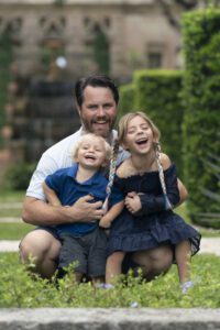 A man crouches outdoors with a young boy and a girl, all smiling and laughing amidst greenery and blurred architectural elements—a perfect moment of family photography Miami style.