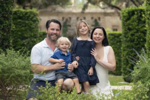 A man, woman, young girl, and young boy pose and smile together outdoors in a lush, green garden setting—perfect for capturing family photography Miami style.