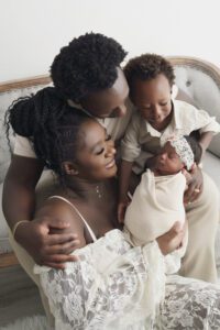 A family of four sits closely together on a couch. Captured by a Miami newborn photographer, the mother holds a swaddled newborn while the father and young boy lean in, all wearing light-colored clothing.