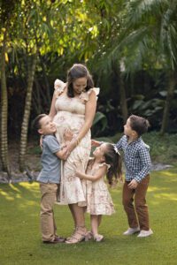 A woman in a floral dress stands on grass, smiling as three young children hug her outdoors with trees in the background.