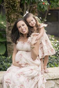 A pregnant woman sits on a stone ledge, smiling, with a young girl standing behind her with an arm around her shoulder. Both wear matching floral dresses. Greenery is in the background.