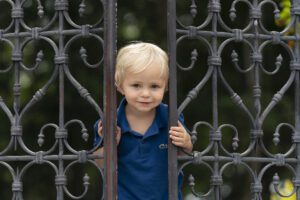 A young child with blond hair in a blue shirt stands behind an ornate metal gate, peering through the bars.