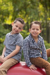 Two young boys with short brown hair sit side by side outdoors, smiling at the camera. One wears a gray polo and khaki pants, the other a plaid shirt and brown pants. Green foliage in the background.
