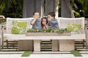 Three young children sit on a white outdoor bench with green leaf pillows, playfully covering their mouths and ears, surrounded by greenery and a succulent planter on the table.