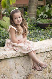 Young girl with long brown hair, wearing a floral dress and sparkly shoes, sits on a stone bench outdoors, smiling at the camera with greenery in the background.