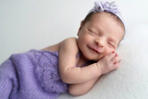 A newborn baby wrapped in a purple blanket with a matching headband smiles while sleeping on a white surface, captured beautifully by a Miami newborn photographer.