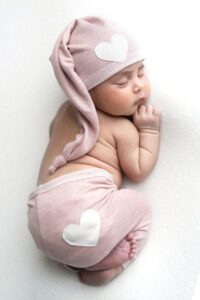 A sleeping baby in pink clothes with white heart patches on the hat and pants lies on a white surface, perfectly captured by a Miami newborn photographer.