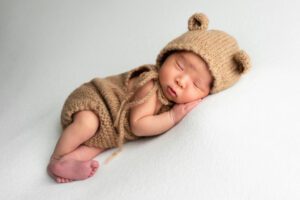Newborn baby sleeping on a white surface, dressed in a brown knitted outfit with a matching bear ear hat—captured beautifully by a Miami newborn photographer.