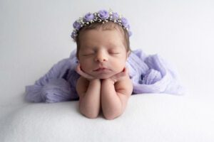 Newborn baby sleeping with chin resting on hands, wearing a purple flower headband, and draped in a light purple blanket against a white background—captured by a Miami newborn photographer.