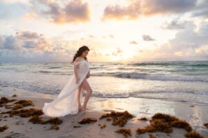 A woman in a flowing white dress stands barefoot on a beach at sunset, with seaweed on the sand and waves in the background.