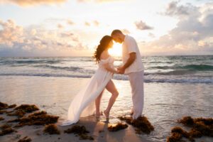 A couple stands barefoot on a beach at sunset, holding hands and touching foreheads, with waves and seaweed in the background.