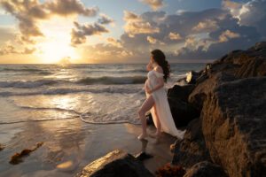 A pregnant woman in a white dress stands by rocky shore at sunset, gazing at the ocean with one hand on her belly.