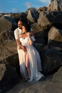 A couple dressed in white stands close together among large rocks at the shoreline, with the woman in a flowing dress and the man embracing her from behind.