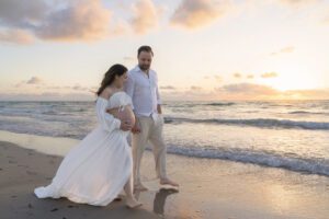 A pregnant woman in a white dress walks barefoot on the beach with a man at sunset, both looking down and smiling.