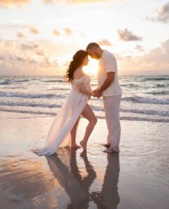 A couple stands face to face holding hands on a beach at sunset, with the woman wearing a flowing white dress and the man in white clothes.