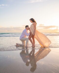 A man kneels on the beach, kissing a pregnant woman’s belly as she stands in a flowing dress, with the ocean and sunset in the background.