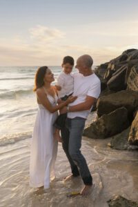 A woman, man, and young boy stand together on a beach near rocks, smiling and interacting as the sun sets over the ocean—a perfect moment for family photography Miami style.