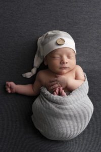 Newborn baby wrapped in a gray blanket, wearing a white hat with a button, peacefully sleeping on a dark gray surface—captured by a Miami newborn photographer.