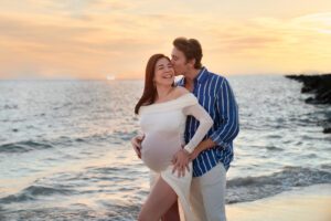 A pregnant woman in a white dress stands on a beach at sunset, smiling, while a man in a blue striped shirt stands behind her and kisses her head.
