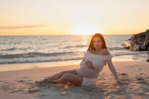 A pregnant woman in a white dress sits on the sand at the beach during sunset, with waves and rocks visible in the background.