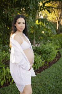 A pregnant woman in a white bra and open shirt stands outdoors in a garden, holding her belly and looking at the camera.