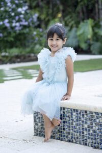 A young girl in a light blue ruffled dress sits barefoot on the edge of a tiled pool, smiling, with greenery and flowers in the background.
