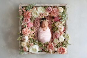 A newborn baby wrapped in a pink blanket lies in a wooden box surrounded by assorted pink and white flowers, wearing a floral headband—capturing the sweet moment as a Miami newborn photographer would.