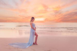 A person in a flowing blue dress stands on a beach at sunset, looking at the ocean.