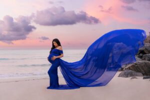 Pregnant woman in a flowing blue dress stands on a beach at sunset, with pink and purple clouds in the sky.