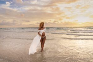 Pregnant woman in a white dress stands on a beach at sunset, holding her belly and looking down at the water.