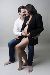A man seated barefoot embraces a barefoot pregnant woman in a black jacket as they look at each other and touch her bare belly in a studio setting.