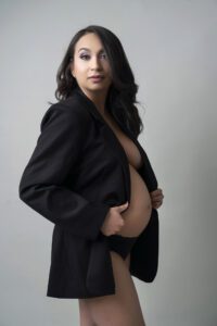 A pregnant woman stands against a plain background, wearing a black blazer and black underwear, holding her blazer open to reveal her baby bump.