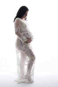 A pregnant woman in a sheer, white lace dress stands against a white background, gently holding her belly and looking down.