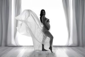 A pregnant woman stands in profile near a window, draped in flowing sheer fabric, with soft natural light illuminating the scene.