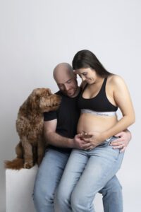 A pregnant woman sits on a man's lap, both holding her belly, while a brown dog sits beside them looking at her. All three appear calm and connected.
