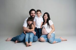 A family of four, including a pregnant woman, man, young boy, and girl, sit barefoot on the floor against a gray background, all wearing casual jeans and white tops.