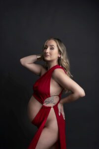 Pregnant woman in a red dress poses against a dark background, with a tattoo visible on her side.