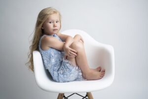 A little girl captured in childhood portraits sitting on a white chair.